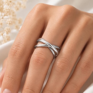 May include: A polished silver-tone ring featuring a crossover design with three smooth, rounded bands that intersect at the centre. The metal has a reflective, high-shine finish, creating a continuous, layered appearance across the finger.