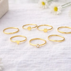 May include: A collection of seven thin gold-toned rings arranged on a white fabric surface. The set includes plain bands, rings with small geometric shapes like a triangle, square, and circle, and one ring featuring a single small clear stone.