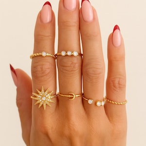 Puede incluir: Una mano que muestra varios anillos de oro con piedras preciosas transparentes. Los anillos incluyen un diseño de estrella, una media luna y bandas con pequeñas piedras. Las uñas están pintadas con una manicura francesa con puntas rojas.