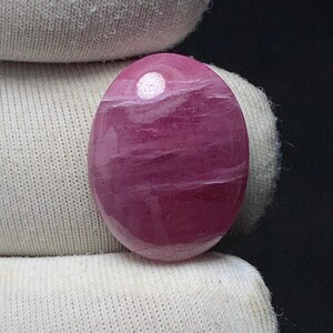 May include: A polished, oval-shaped gemstone in shades of pink and magenta. The stone has a smooth, glossy surface with subtle white veining. The gemstone is held against a white fabric background, with a dark background.