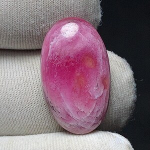 May include: A polished, oval-shaped gemstone in shades of pink and white. The stone has a smooth, glossy surface with a marbled appearance. The gemstone is held in a white fabric.