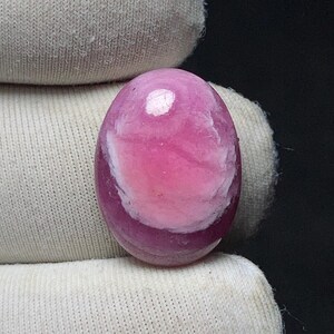 May include: A polished, oval-shaped gemstone with a vibrant pink hue, featuring a swirling pattern of lighter pink and white. The gemstone is held against a neutral background, highlighting its color and texture.