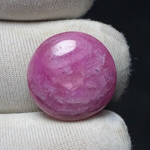 May include: A polished, round, pink gemstone with a translucent appearance. The gemstone has a smooth surface with subtle internal patterns and a slight sheen. The gem is held against a white fabric background.