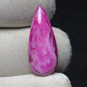 May include: A polished, teardrop-shaped gemstone. The stone displays a mix of pink and white colours, giving a marbled appearance. The gemstone is set against a white fabric background.