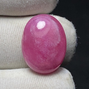 May include: A polished, oval-shaped, pink gemstone. The stone has a smooth, glossy surface with a lighter, almost white, spot near the top. The gemstone is held by a white fabric.