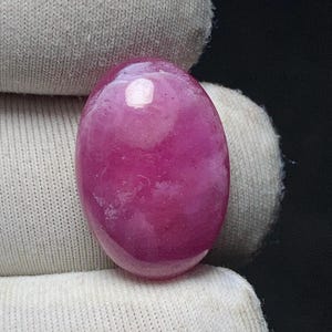 May include: A polished, oval-shaped gemstone in a vibrant magenta hue. The stone has a smooth, glossy surface with subtle internal variations, resting on a white fabric background. The gemstone's color is rich and saturated, with a slight translucence.