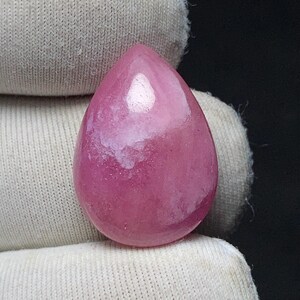 May include: A polished, teardrop-shaped gemstone in shades of pink and white. The stone has a smooth, glossy surface and is held against a white fabric background. The gemstone's color is a vibrant pink with white marbling.
