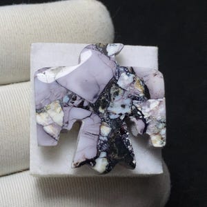 May include: A handcrafted stone carving in the shape of a bird, featuring a mix of lavender, black, white, and gray colors. The bird is mounted on a square, off-white base. The stone has a unique, marbled appearance.