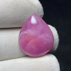 May include: A polished, teardrop-shaped gemstone in shades of pink. The gem has a smooth, glossy surface and is held by a white fabric. The gem's color varies from light to deep pink, with subtle internal patterns.