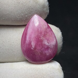 May include: A polished, teardrop-shaped gemstone in shades of pink and magenta. The stone has a smooth, glossy surface with subtle white veining. The gemstone is held against a white fabric background.
