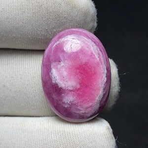 May include: Oval-shaped gemstone with a vibrant pink hue, featuring a swirling pattern of white and darker pink. The polished stone is held against a white fabric background, showcasing its smooth texture and color variations. A close-up view highlights the gem's natural beauty.