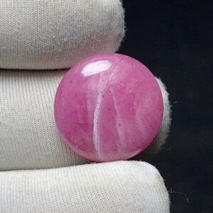 May include: A round, polished gemstone in a vibrant pink hue, with white veining. The stone is held by a white fabric. The gemstone's surface reflects light, highlighting its smooth texture and color variations.