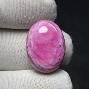 May include: A polished, oval-shaped gemstone in shades of pink and white. The stone has a swirling pattern and a glossy finish. The gemstone is set against a white fabric background.