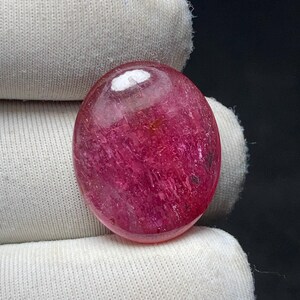 May include: A polished, oval-shaped gemstone with a deep pink hue. The stone has a translucent quality, with visible internal structures and a glossy surface. The gem is set against a white fabric background, highlighting its color and form.