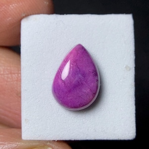 May include: A polished, teardrop-shaped gemstone in shades of deep purple. The stone has a smooth, glossy surface with internal marbling, set against a white background. The gem is likely used for jewelry or decorative purposes.