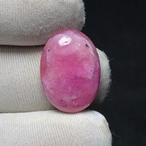 May include: A polished, oval-shaped gemstone in shades of pink. The stone has a smooth, glossy surface with subtle variations in color and texture. The gemstone is held against a white fabric background.