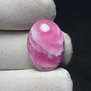 May include: Oval-shaped pink gemstone with white swirling patterns. The polished stone has a smooth surface and a prominent white spot near the top. The gemstone is held against a white fabric background.