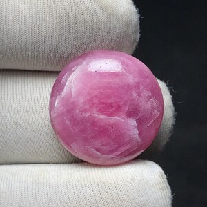 May include: A polished, round, pink gemstone. The stone has a smooth, reflective surface with subtle variations in color and texture. The gemstone is held by a white fabric.