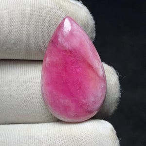 May include: A polished, teardrop-shaped gemstone in shades of pink and magenta. The stone has a translucent quality with swirling patterns and a glossy finish. The gem is held against a white fabric background.