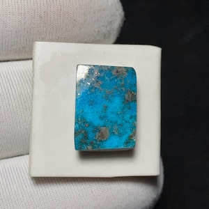 Pode incluir: Uma pedra preciosa retangular de turquesa com superfície polida. A pedra apresenta um tom azul vibrante com inclusões cinza escuro e prateadas. Está colocada sobre uma base branca retangular. Ideal para a criação de joias.