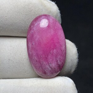 May include: A polished, oval-shaped gemstone in a vibrant magenta hue. The stone has a smooth, reflective surface with subtle internal veining and a bright, circular highlight. The gem is set against a white fabric background.