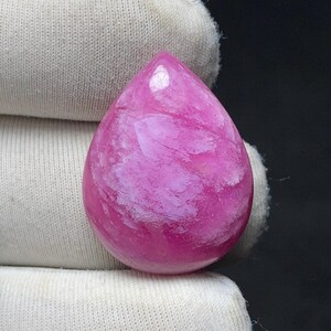 May include: A polished, teardrop-shaped gemstone in shades of pink and white. The stone has a glossy finish and a marbled appearance, with swirling patterns of lighter pink and white throughout. The gem is held against a white fabric background.