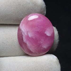 May include: A polished, oval-shaped gemstone with a vibrant pink hue, featuring swirling white inclusions. The stone is held against a white fabric background, showcasing its smooth, reflective surface. This gemstone is ideal for jewelry making.