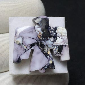 May include: A handcrafted stone carving in the shape of a bird, featuring a mix of colors including lavender, black, white, and tan. The bird carving is set on a white surface, showcasing the intricate details of the stone.