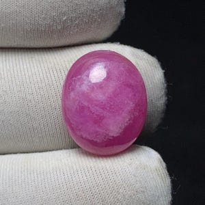 May include: A polished, oval-shaped gemstone in shades of pink and white. The stone has a glossy finish and a marbled appearance, with swirling patterns of white within the pink base. The gemstone is set against a white fabric background.