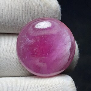 May include: A polished, round, ruby gemstone with a vibrant pinkish-red hue. The stone has a smooth, reflective surface with internal swirls and a white inclusion. The gemstone is set against a white fabric background.