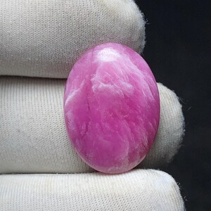 May include: A polished, oval-shaped gemstone in a vibrant pink hue, with swirling white inclusions. The stone is smooth and appears to be held by a white fabric. The gemstone's shape and color suggest it could be used in jewelry or as a decorative element.