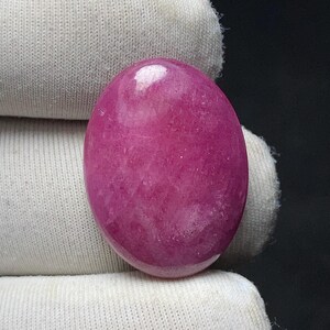 May include: A polished, oval-shaped gemstone in a vibrant magenta hue. The stone has a smooth, glossy surface and a slightly translucent appearance. The gemstone is set against a white fabric background.
