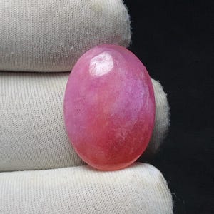 May include: A polished, oval-shaped gemstone in shades of pink and red. The stone has a smooth, glossy surface and appears to be held by a white fabric. The gemstone is a vibrant and eye-catching piece.