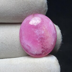May include: A polished, oval-shaped gemstone in shades of pink and white. The stone has a smooth, glossy surface with a marbled appearance. The gemstone is set against a white fabric background, highlighting its color and texture.