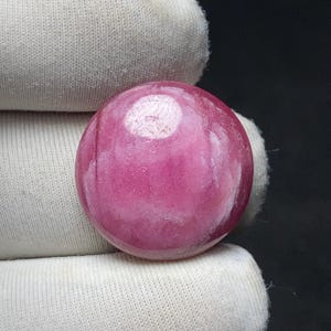 May include: A polished, spherical gemstone in a vibrant pink hue. The surface has subtle variations in color and texture, with a prominent white area. The gemstone is held against a white fabric background, creating a contrast that highlights its color and form.