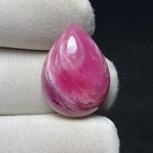 May include: A polished, teardrop-shaped gemstone in shades of pink and white. The stone has a smooth, glossy surface with swirling patterns. The gem is held by a white fabric.