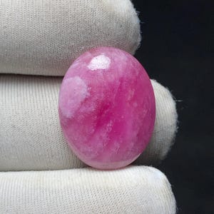 May include: A polished, oval-shaped gemstone in shades of pink and white. The stone has a smooth, glossy surface with a marbled appearance. The gemstone is held in a white fabric.