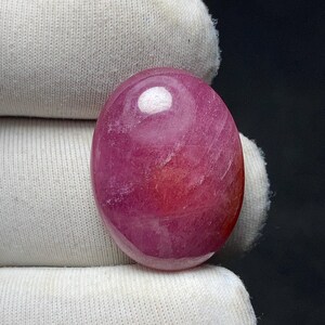 May include: A polished, oval-shaped gemstone in shades of pink and red. The stone has a smooth, glossy surface with a prominent white reflection. The gemstone is held against a white fabric background.