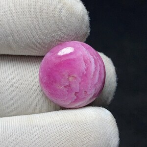 May include: A polished, round gemstone in shades of pink and white. The stone has a swirling pattern and a glossy finish. The gemstone is held by a white fabric.