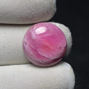 May include: A polished, round gemstone with a vibrant pink hue and swirling white patterns. The gemstone is held in a white fabric, showcasing its smooth, reflective surface. The gem's color ranges from deep rose to lighter pink.