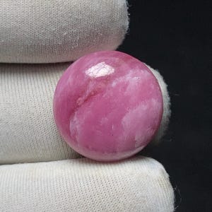 May include: A polished, round gemstone in shades of pink and white. The stone has a smooth, glossy surface and appears to be held by a white fabric. The gemstone is a sphere, and the colors blend together.