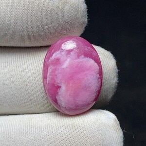 May include: A polished, oval-shaped gemstone with a vibrant pink hue and a cloudy white pattern. The stone is smooth and reflective, with a glossy finish. The gemstone is held in a white fabric.