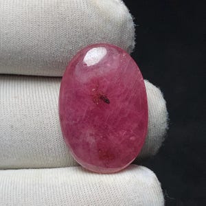 May include: A polished, oval-shaped gemstone in a deep pink hue. The stone has a smooth surface with internal inclusions and a small, dark spot. The gemstone is set against a white fabric background.