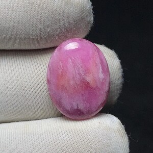 May include: A polished, oval-shaped gemstone with a swirling pattern of pink and white hues. The stone is smooth and glossy, with variations in color creating a marbled effect. The gemstone is set against a white fabric background.