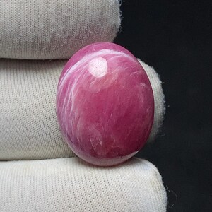 May include: A polished, oval-shaped gemstone in shades of pink and white. The stone has a smooth, glossy surface with swirling patterns of white against a deep pink background. The gemstone is held by a white fabric.