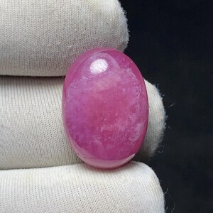 May include: A polished, oval-shaped gemstone in a vibrant shade of pink. The stone has a smooth, glossy surface with subtle internal patterns. The gem is held against a white fabric background, with a dark backdrop.
