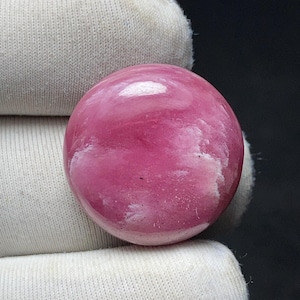 May include: A polished, oval-shaped gemstone with a vibrant pink hue, featuring swirling white inclusions. The stone's surface reflects light, creating a glossy appearance. The gemstone is set against a white fabric background.