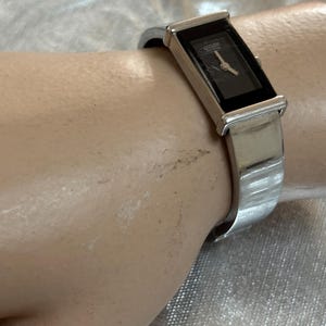 May include: A silver-toned Citizen watch with a rectangular black face and a silver band. The watch has a minimalist design with a sleek, modern aesthetic. The watch is worn on a wrist.