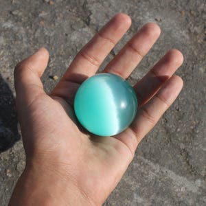 Puede incluir: Una esfera redonda de color turquesa con un efecto de ojo de gato blanco, sostenida en una mano abierta. La esfera es lisa y translúcida, con una superficie brillante y reflectante. El fondo es una superficie rugosa y gris.