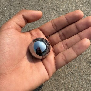 May include: A small, highly polished metal sphere with a reflective surface. The sphere reflects the surrounding environment. It is held in an open hand, palm upwards.
